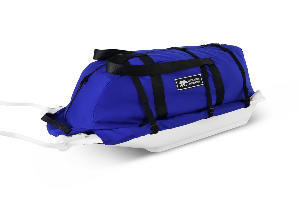 A Royal Blue snowmobile Sled Pulk cargo bag with black straps and a white plastic base, shown on a white studio background. With a tag labeled 'Wilderness Engineering'.