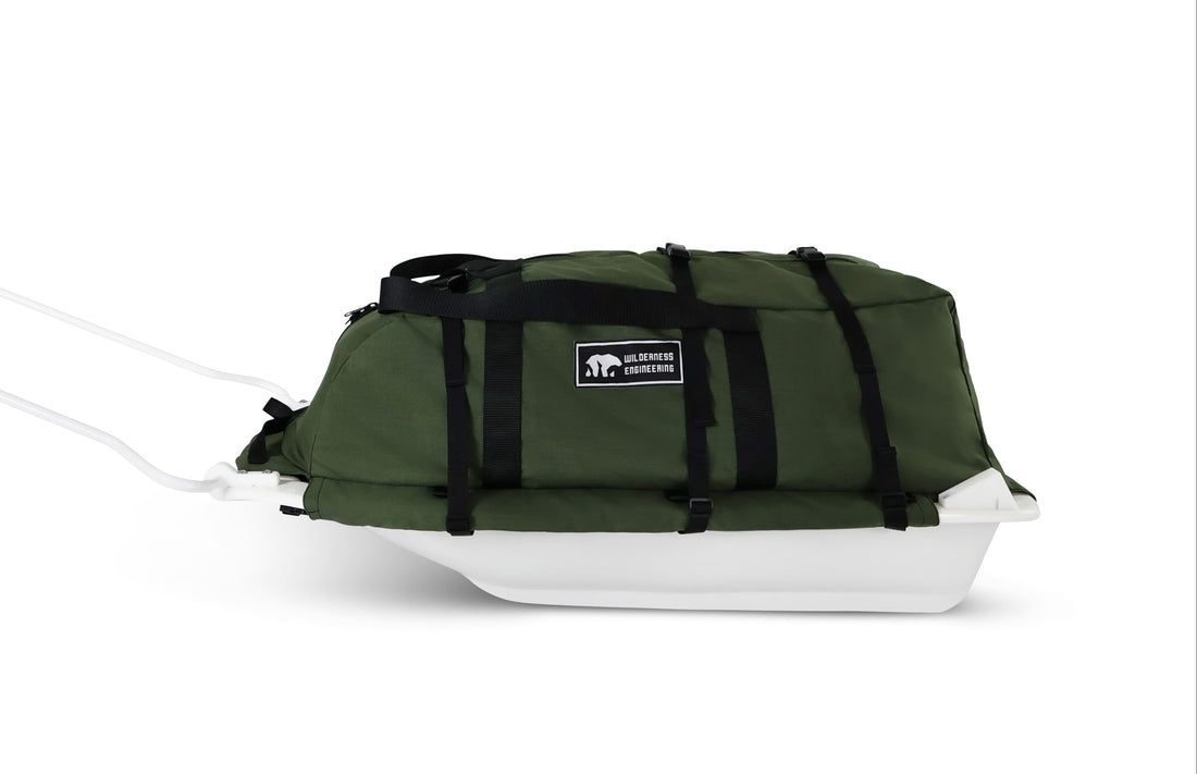 A Olive Green snowmobile Sled Pulk cargo bag with black straps and a white plastic base, shown on a white studio background. With a tag labeled 'Wilderness Engineering'.