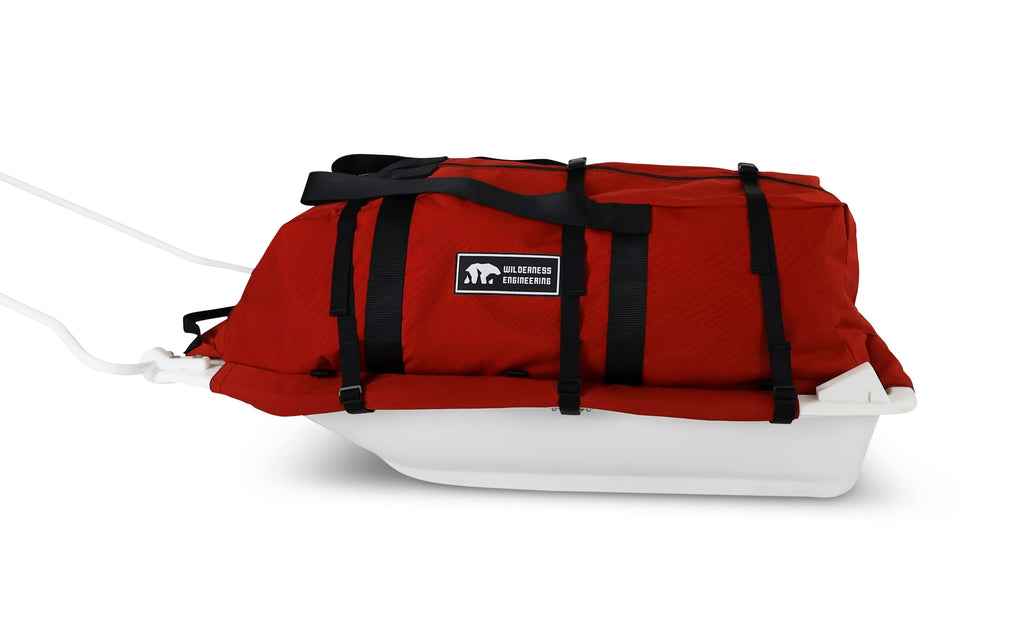 A Red snowmobile Sled Pulk cargo bag with black straps and a white plastic base, shown on a white studio background. With a tag labeled 'Wilderness Engineering'.