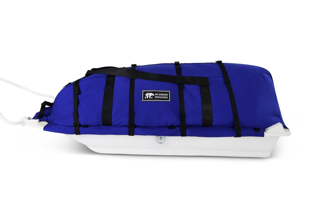 A blue snowmobile pulk sled cargo bag with black straps and a white plastic sled base, shown on a clean white background. A black label on the front displays the brand 'Wilderness Engineering'.