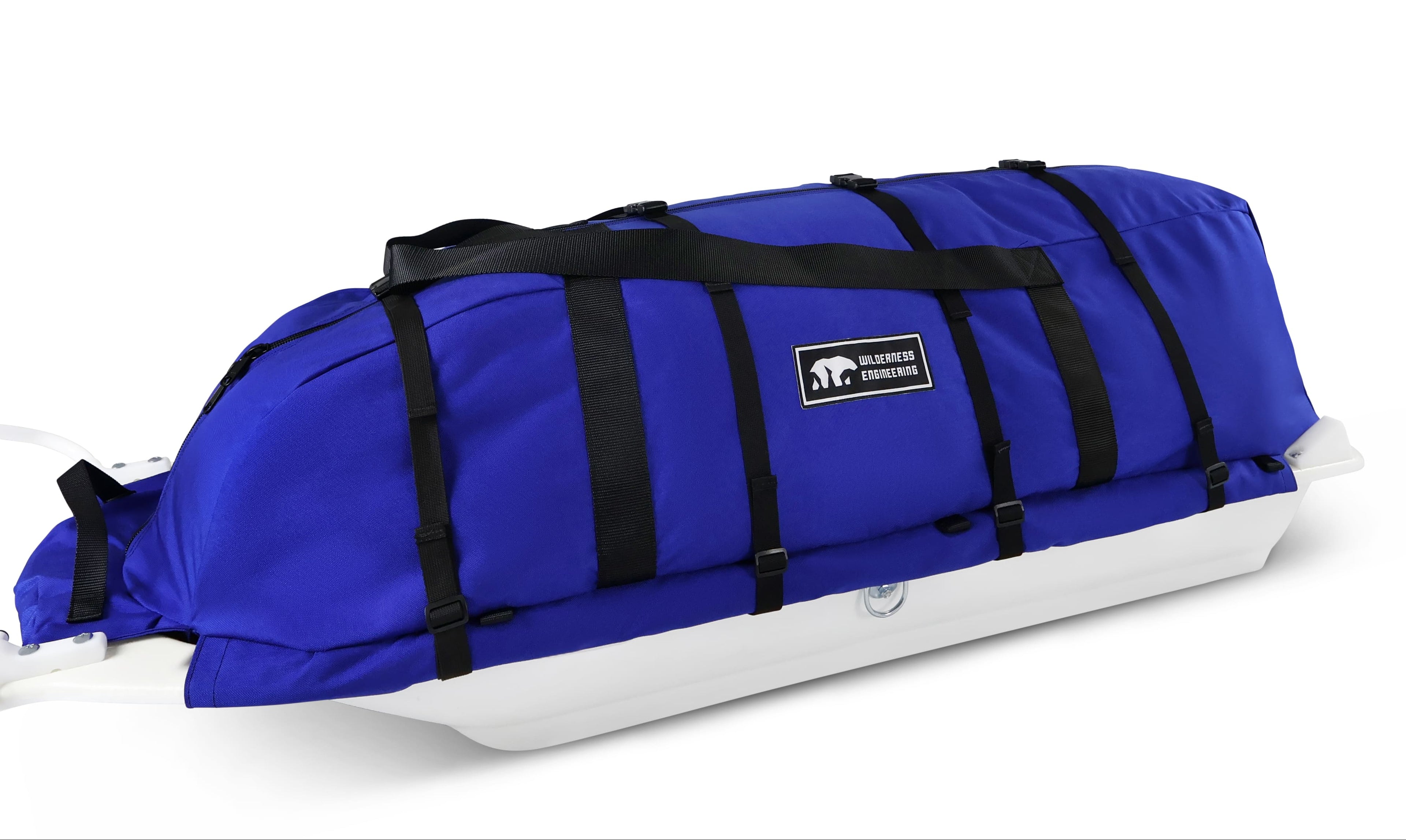A blue snowmobile pulk sled cargo bag with black straps and a white plastic sled base, shown on a clean white background. A black label on the front displays the brand 'Wilderness Engineering'.