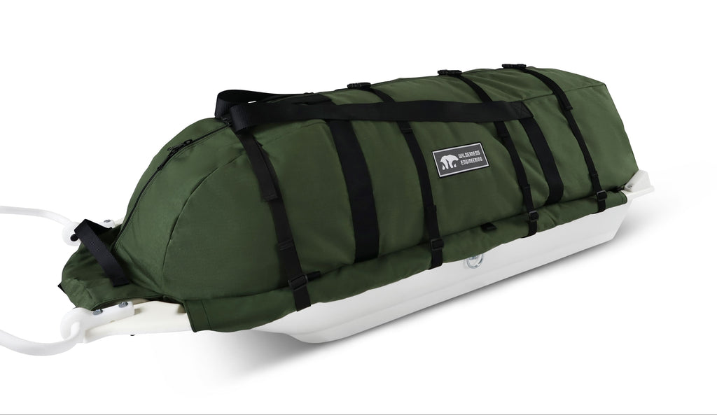 An olive green snowmobile cargo bag with black straps and a white plastic base, shown on a white studio background. With a tag labeled 'Wilderness Engineering'.