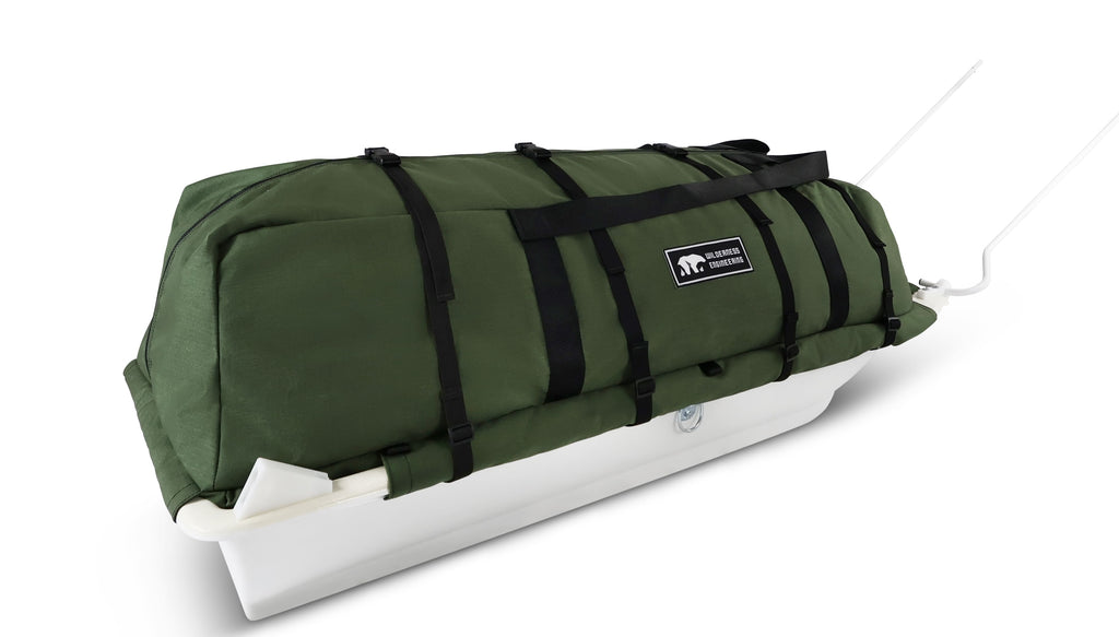 An olive green snowmobile cargo bag with black straps and a white plastic base, shown on a white studio background. With a tag labeled 'Wilderness Engineering'.