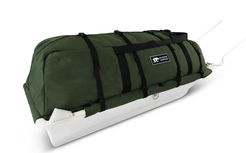 An olive green snowmobile cargo bag with black straps and a white plastic base, shown on a white studio background. With a tag labeled 'Wilderness Engineering'.