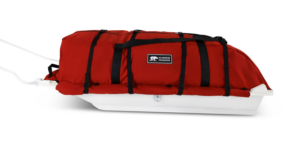 An Red snowmobile sled pulk cargo bag with black straps and a white plastic base, shown on a white studio background. With a tag labeled 'Wilderness Engineering'.