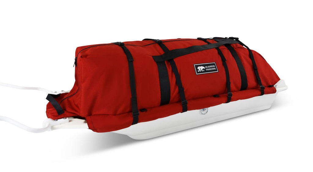 An Red snowmobile Sled Pulk cargo bag with black straps and a white plastic base, shown on a white studio background. With a tag labeled 'Wilderness Engineering'.