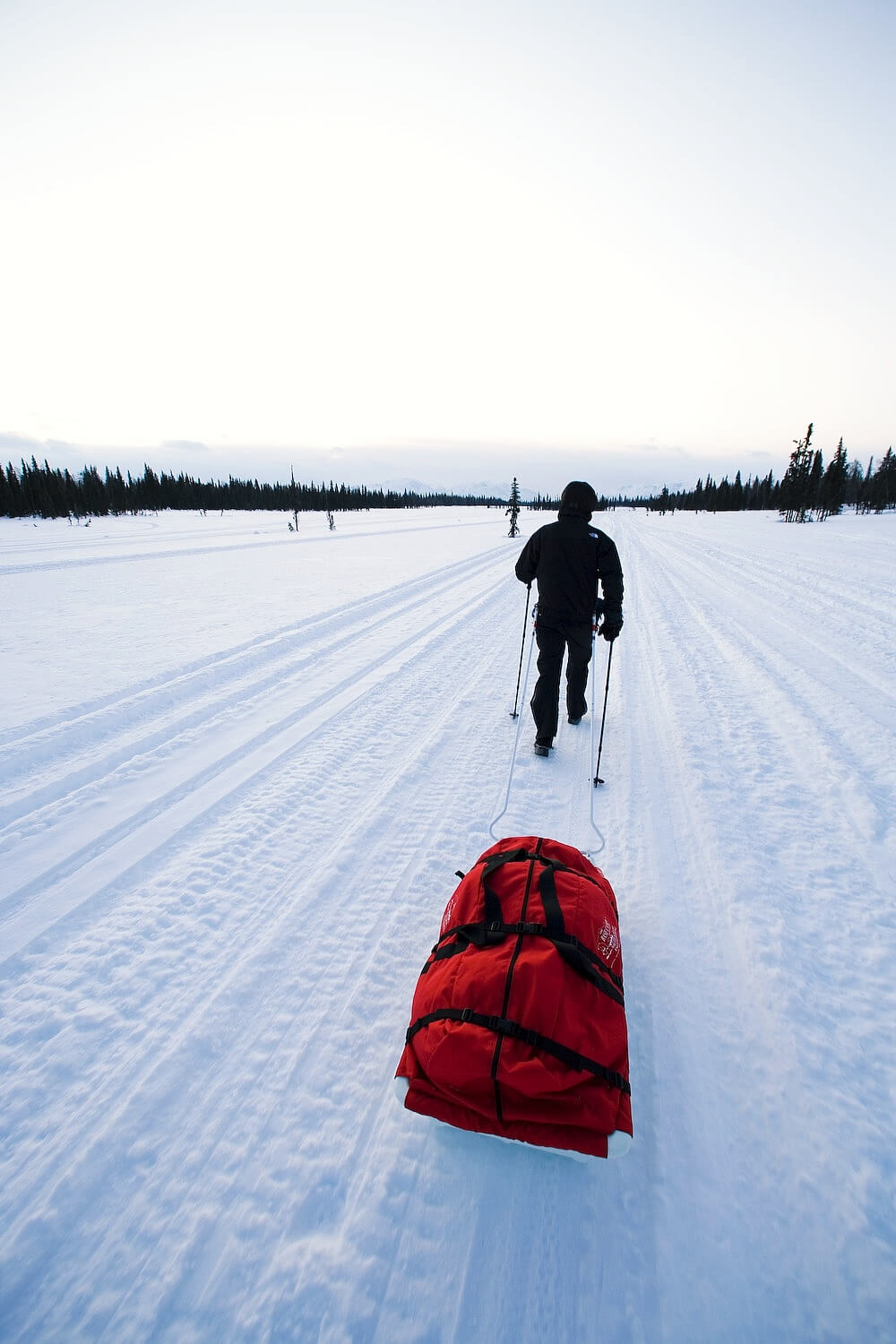 An Red snowmobile Sled Pulk cargo bag with black straps and a white plastic base, shown on a white studio background. With a tag labeled 'Wilderness Engineering'.